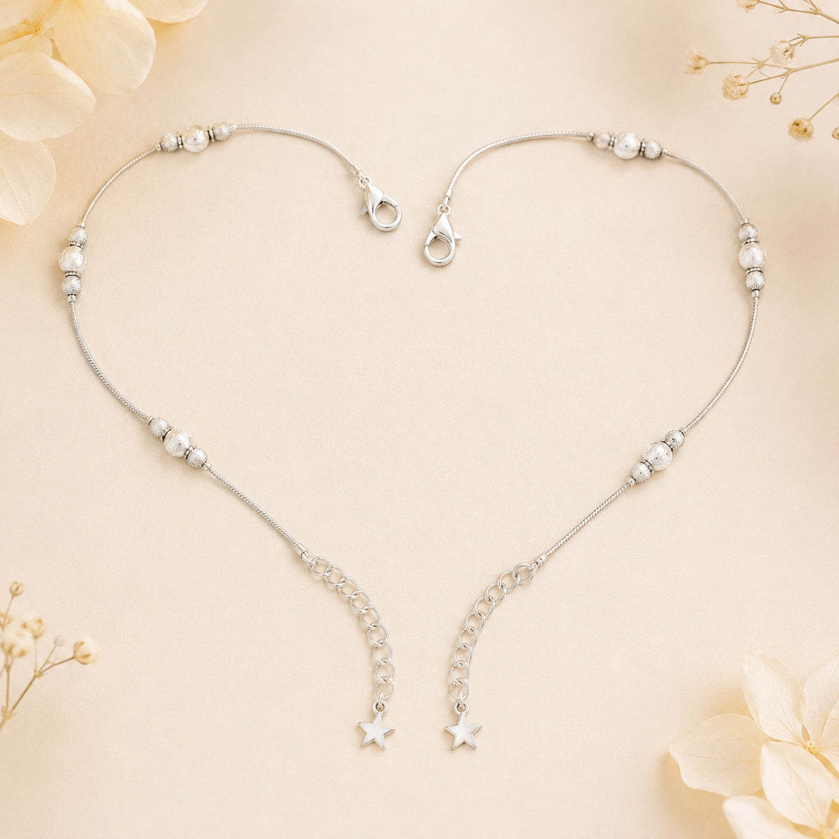 Silver-tone beaded anklets arranged in heart shape on beige surface