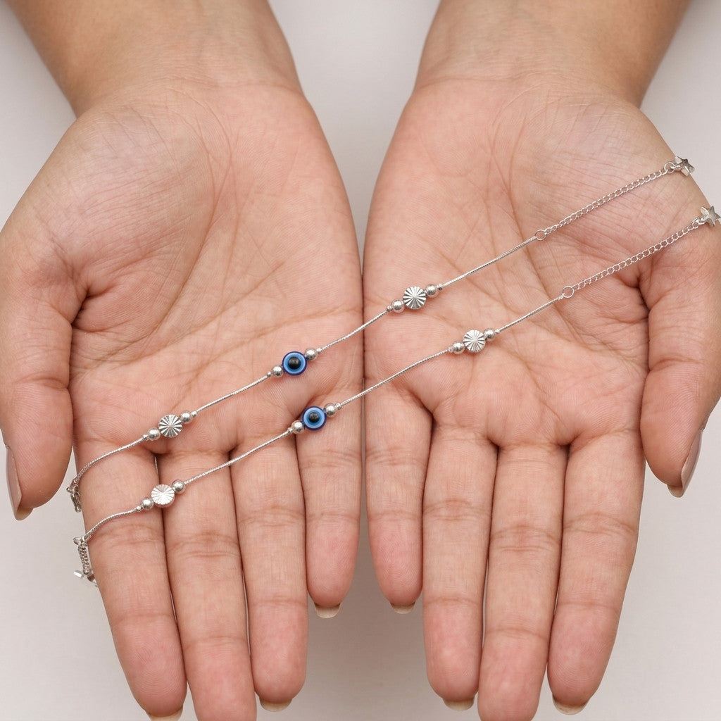 Two silver-tone anklets with blue evil eye charm held in hands